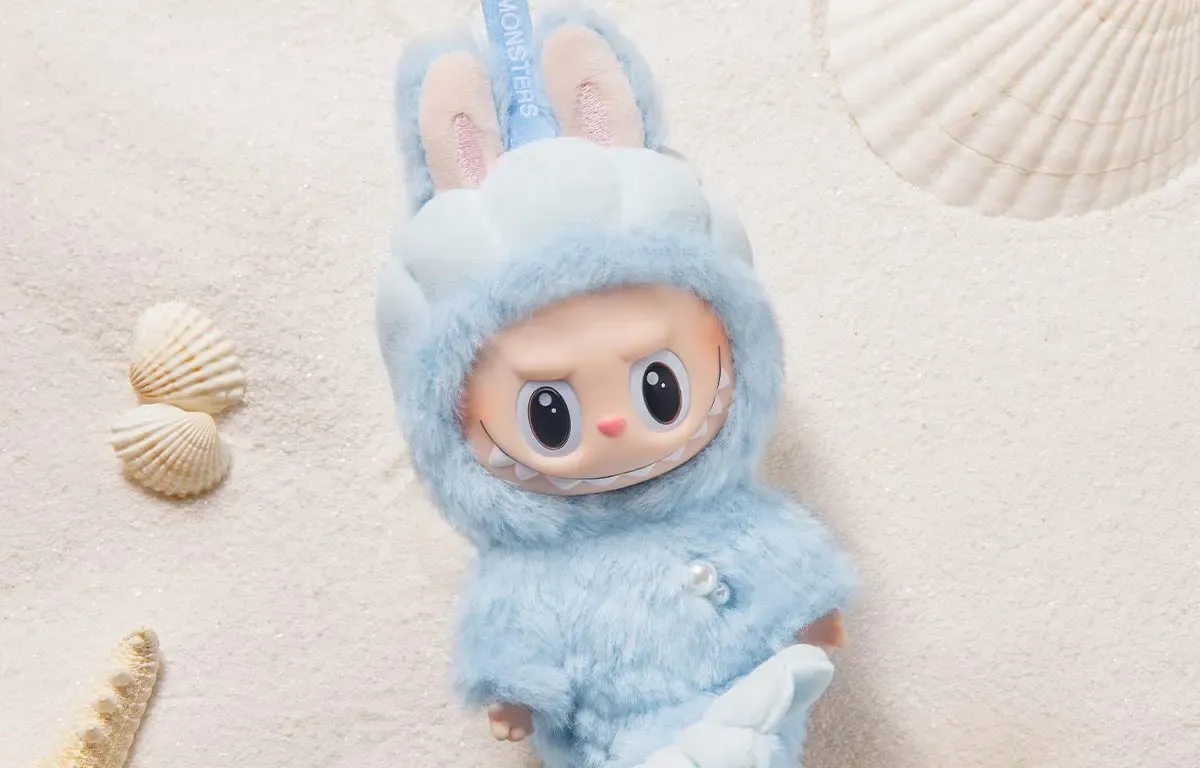 Roztomilá modrá figurka Labubu s králičíma ušima na pískovém pozadí.